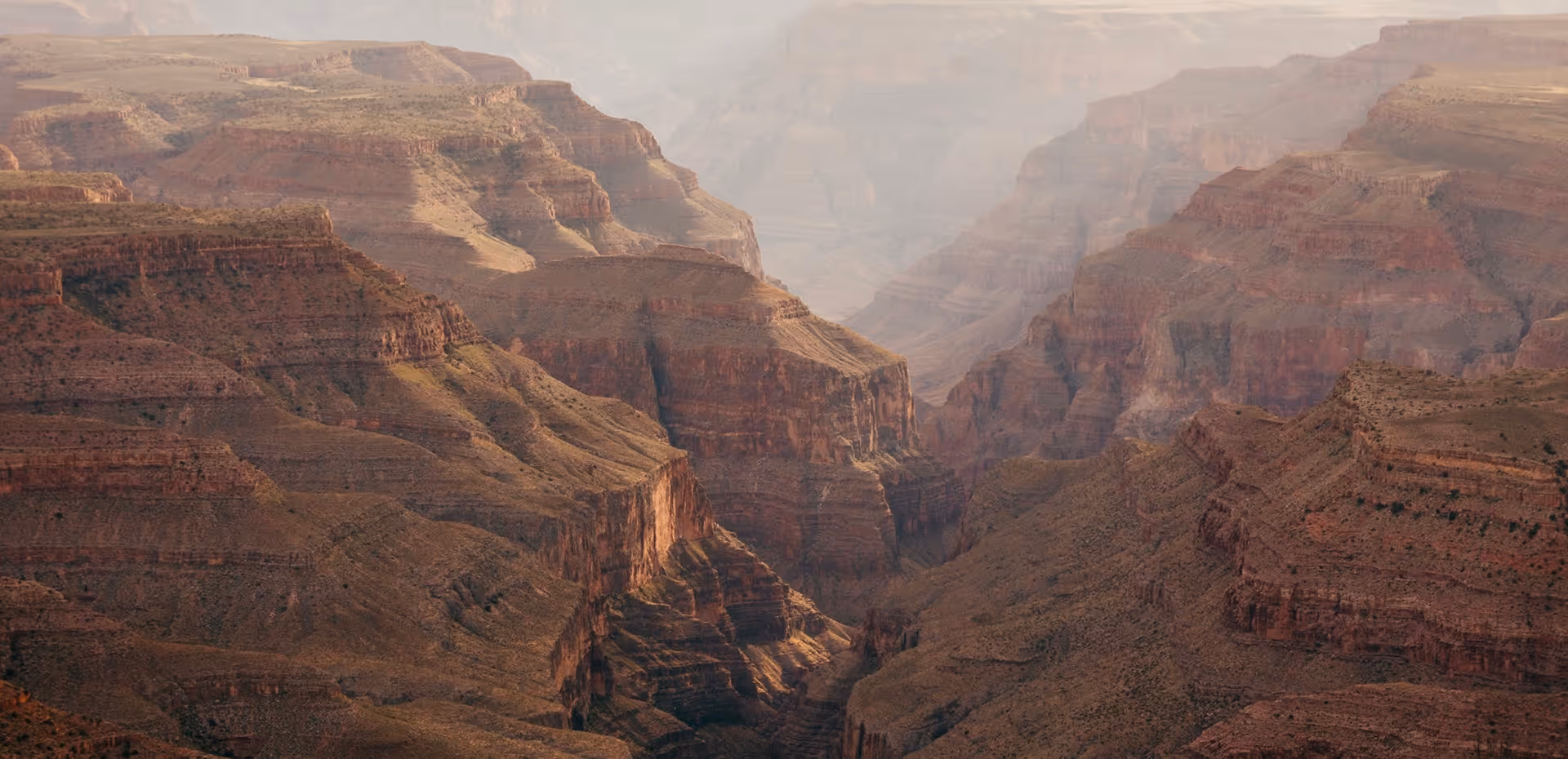 Vast layered rock formations and cliffs of the Grand Canyon under soft sunlight with a hazy atmosphere.